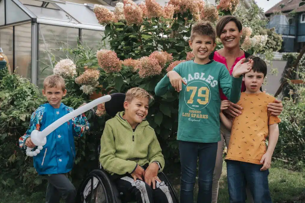 Marina mit vier Kindern aus dem Ferienprogramm: Ein Kind mit Luftballon-Schwert, ein lächelnder Junge im Rollstuhl, ein Junge, der in die Kamera winkt, und ein kleiner Junge, den Marina an den Schultern hält.