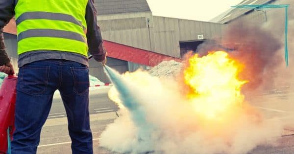 Eine Person in einer gelben Warnweste benutzt einen Feuerlöscher, um ein Feuer zu löschen. Im Hintergrund sind industrielle Gebäude zu sehen. Unten rechts befinden sich die Logos von "Kaspar Arbeitssicherheit und Brandschutz" und "ErsteHilfe Ortenau".