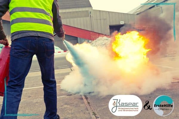 Eine Person in einer gelben Warnweste benutzt einen Feuerlöscher, um ein Feuer zu löschen. Im Hintergrund sind industrielle Gebäude zu sehen. Unten rechts befinden sich die Logos von "Kaspar Arbeitssicherheit und Brandschutz" und "ErsteHilfe Ortenau".