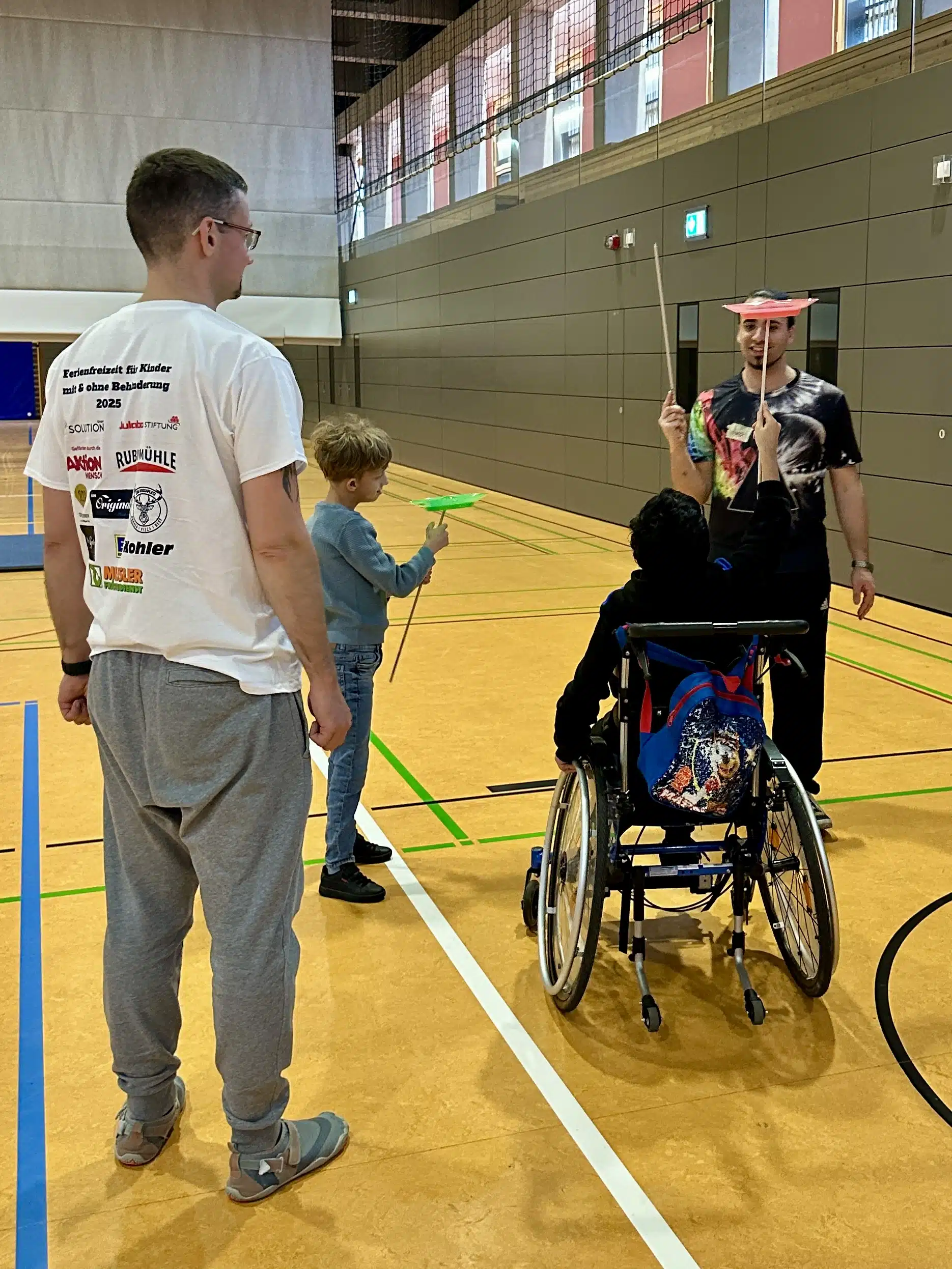 Kinder üben mit Jongliertellern in der Turnhalle, darunter ein Kind im Rollstuhl, während ein Betreuer unterstützt – Aktivität im inklusiven Ferienprogramm Herbst 2025