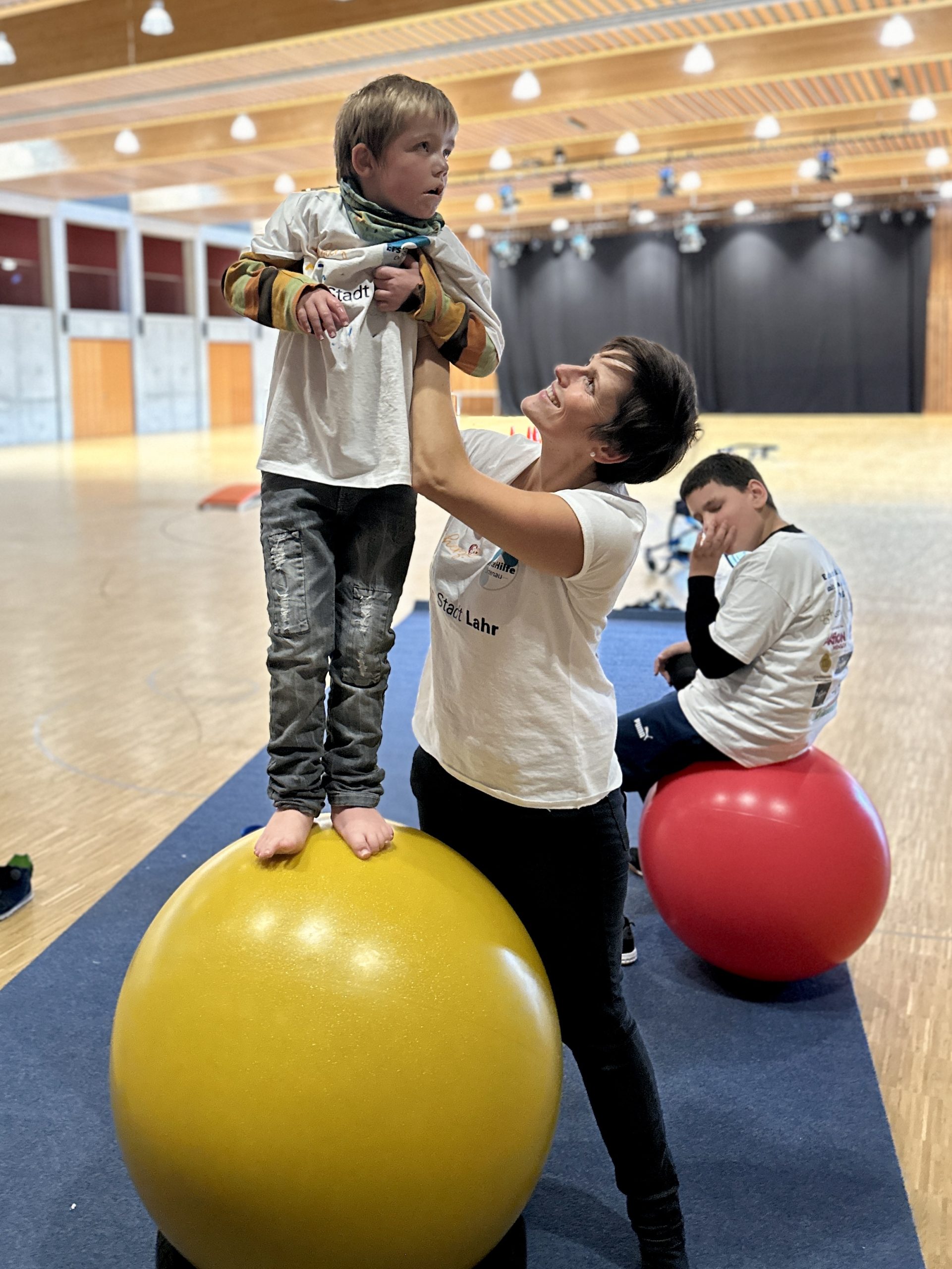 Kind steht auf einem großen gelben Gymnastikball, unterstützt von einer Betreuungsperson, während eine weitere Person auf einem roten Ball sitzt – Bewegungseinheit im inklusiven Ferienprogramm Herbst 2025