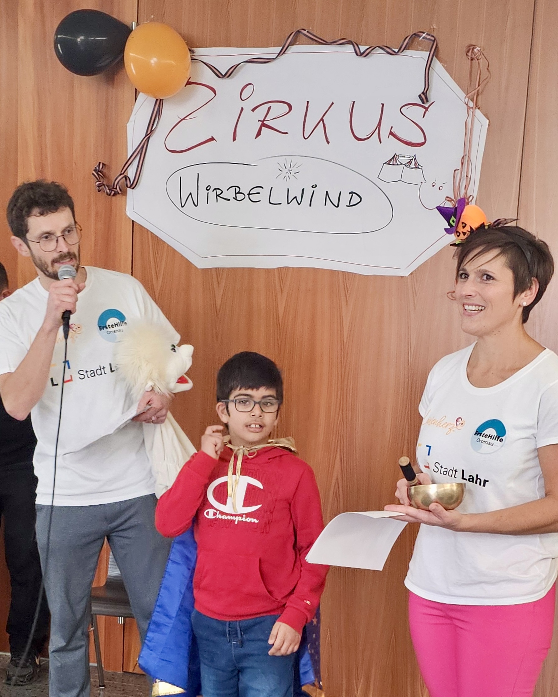 Zwei Betreuungspersonen und ein Kind stehen vor einem Zirkusplakat mit der Aufschrift „Zirkus Wirbelwind“ während der Dankesrede beim Abschlussfest des inklusiven Ferienprogramms Herbst 2025