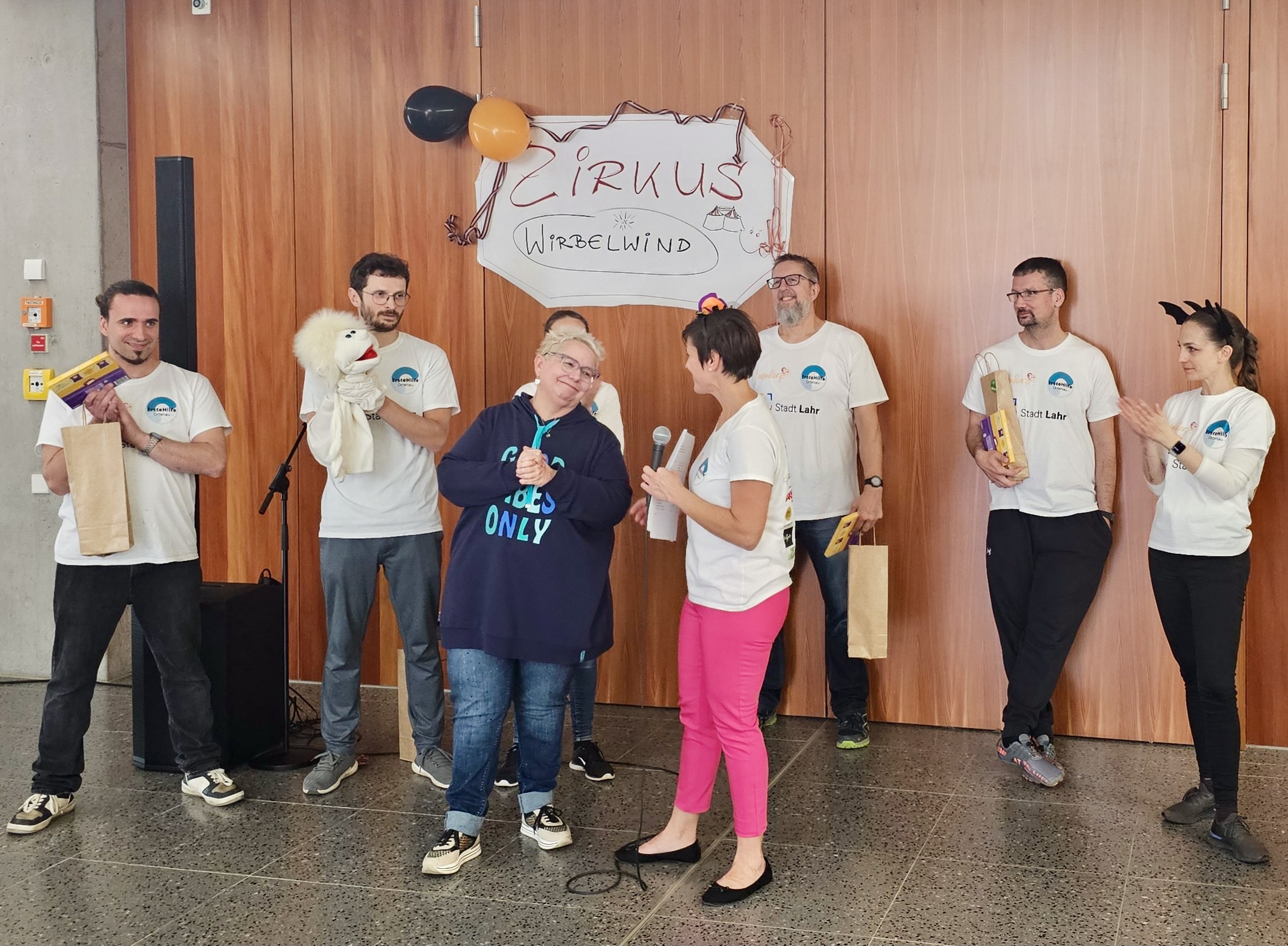 Betreuer stehen vor einem Zirkusplakat „Zirkus Wirbelwind“ und erhalten Geschenke während der Dankesrede beim Abschlussfest des inklusiven Ferienprogramms Herbst 2025